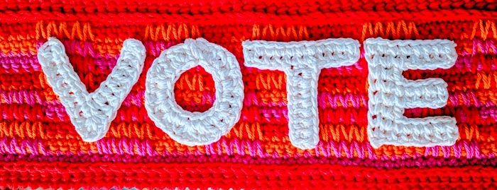 Crocheted letters V-O-T-E against sunrise-colored crochet background