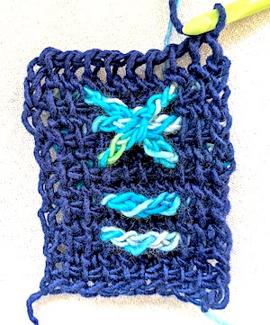 Ten-stitch rows with a solid dark color background of Tunisian extended simple stitch and variegated light blue raised horizontal bars over 4 stitches of the row, and at the top last rows, three of these bars are wrapped with a stitch in the middle to resemble a raised asterisk shape